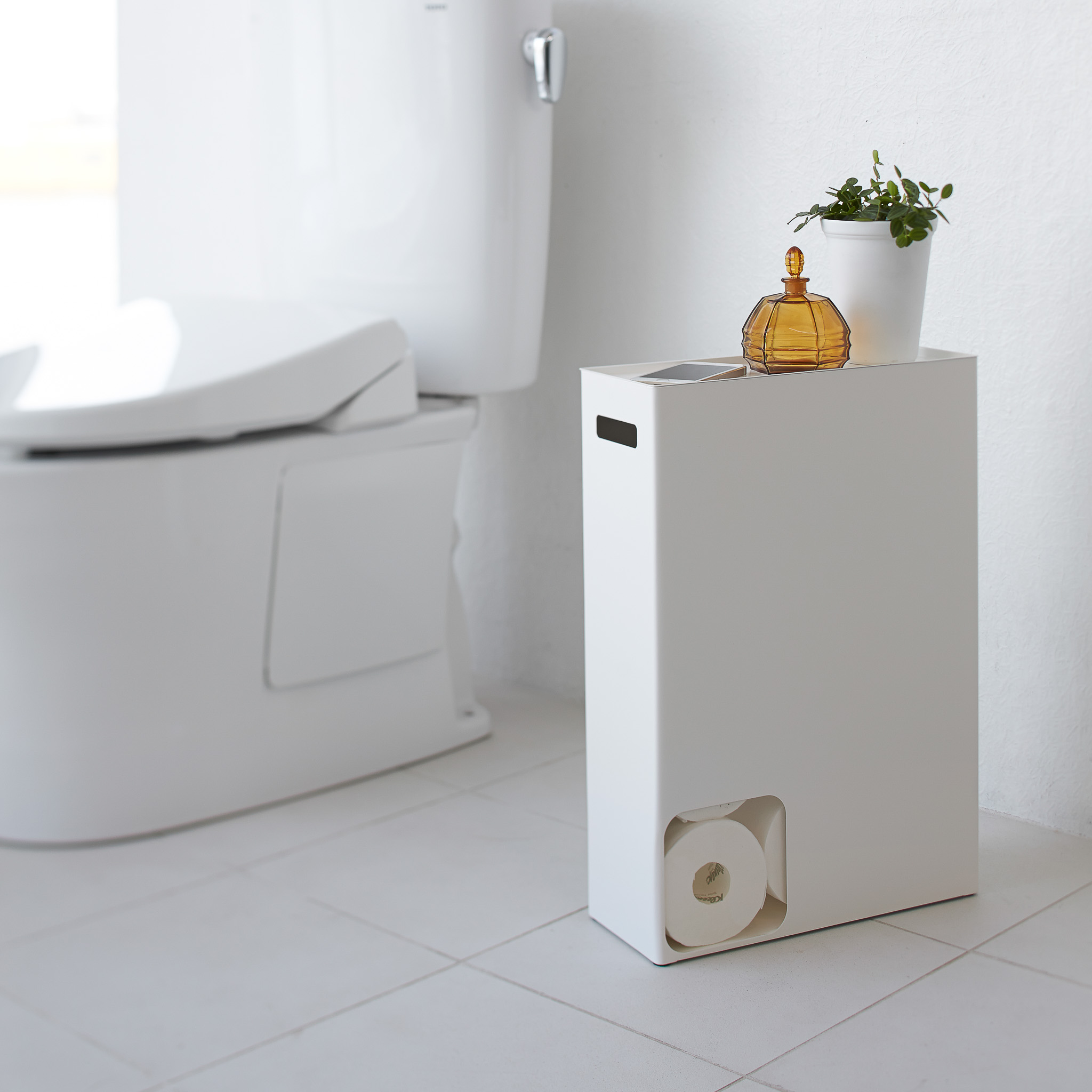 Vue de face d'un stockeur de papier toilette blanc Yamazaki Home contenant du papier toilette et des objets de d?coration dans la salle de bain.