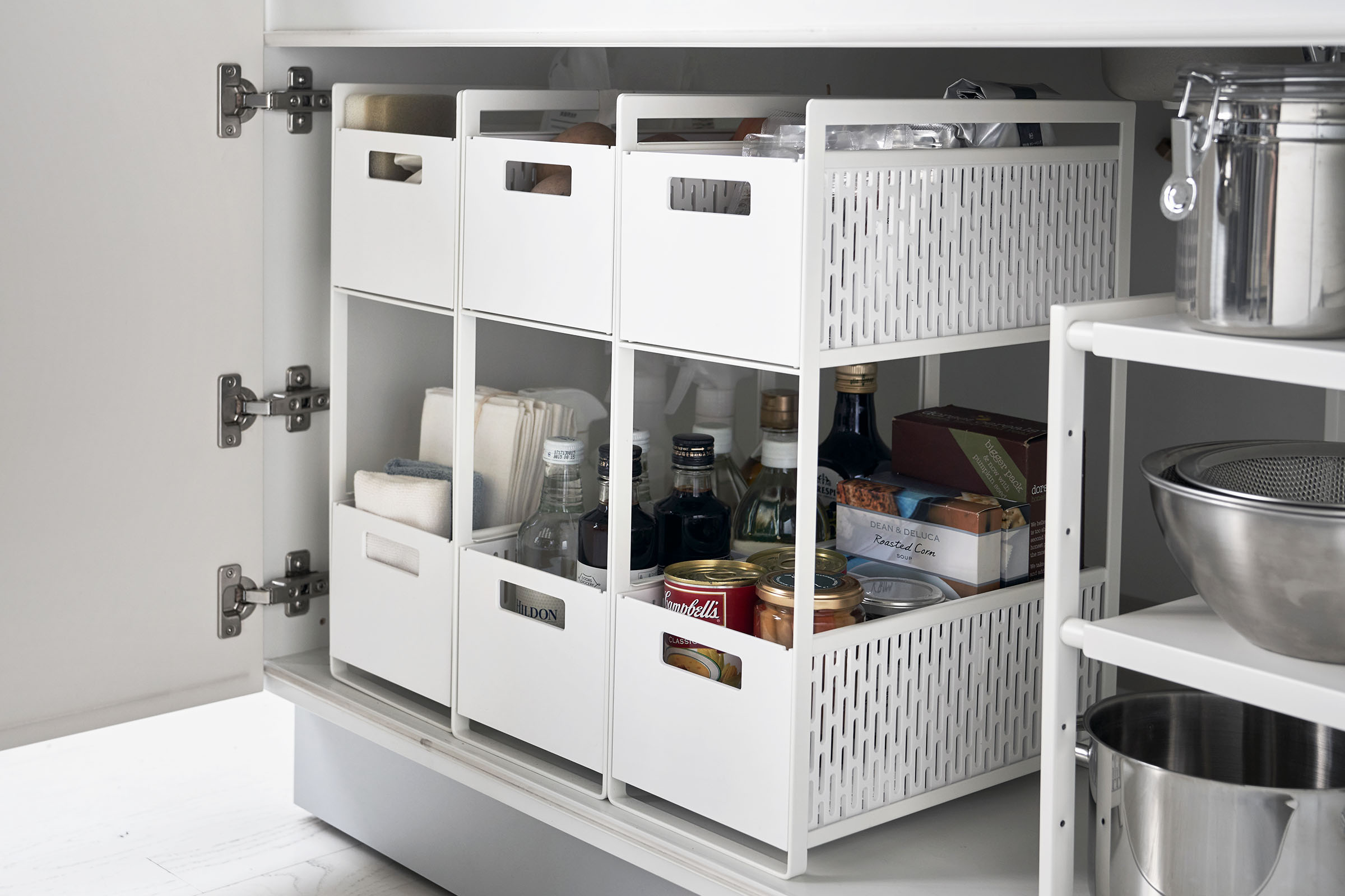 Three two-tier white storage bins are displayed in a lower cabinet. The first houses cleaning supplies, the other two hold pantry items.