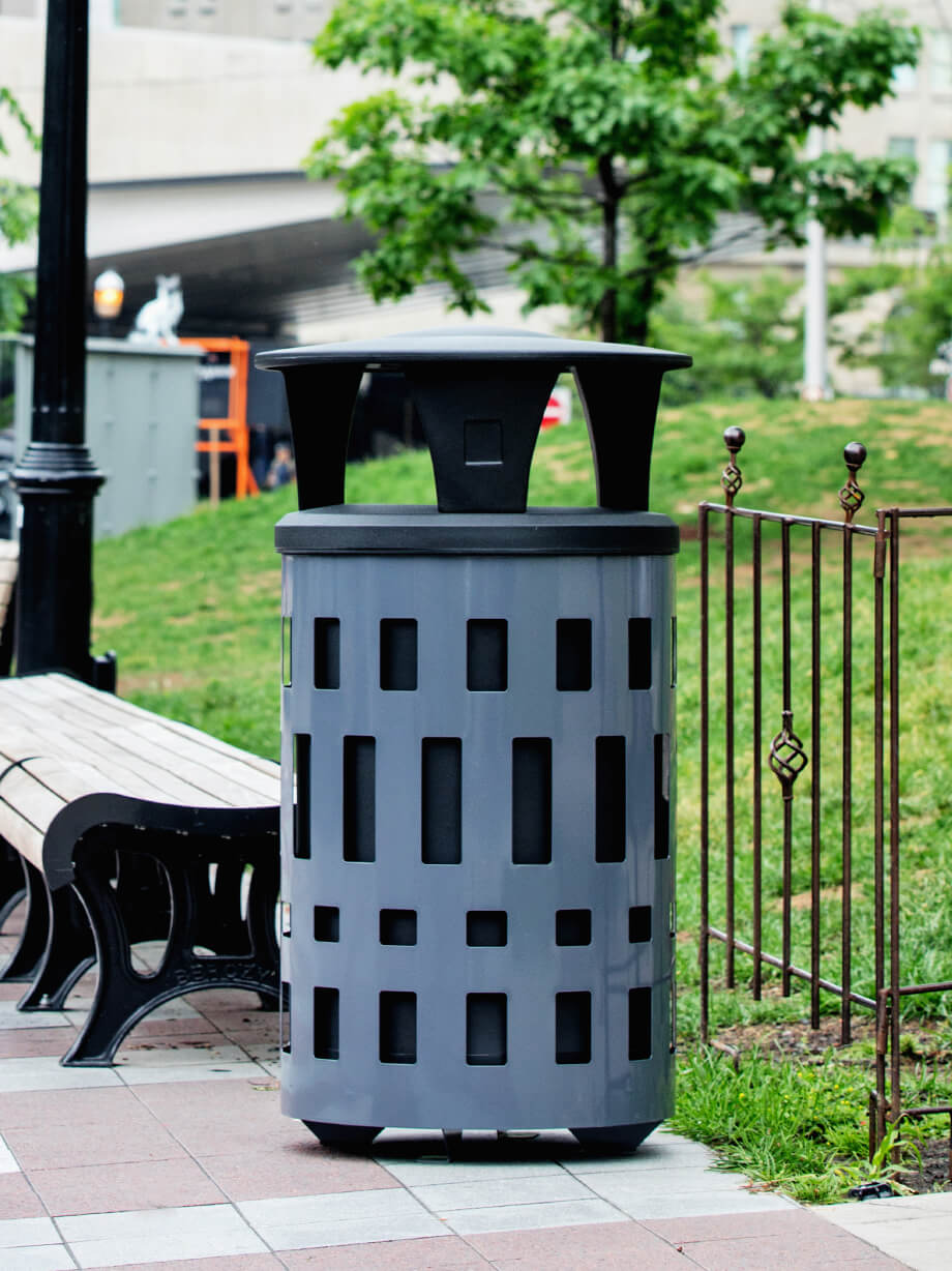 metal outdoor durable waste container with a plastic canopy lid and large capacity liners beside a bench in a city park
