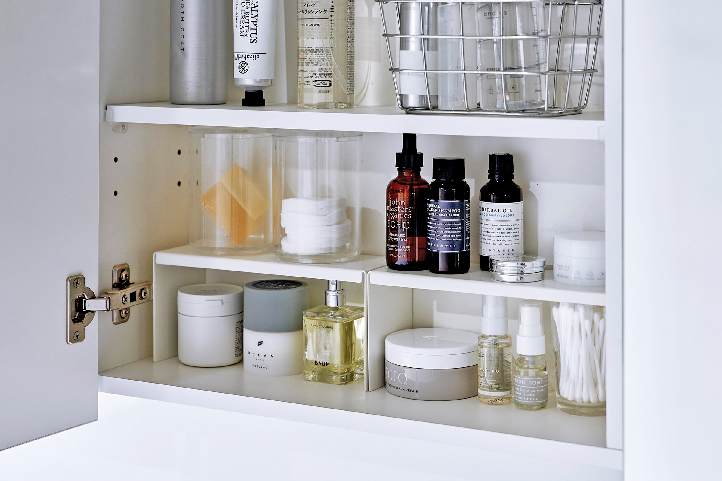 Open medicine cabinet showing two white countertop risers on bottom shelf, side by side. Upper level of the riser holds perfume and hair products, while skincare and small sprays are stored on the bottom.