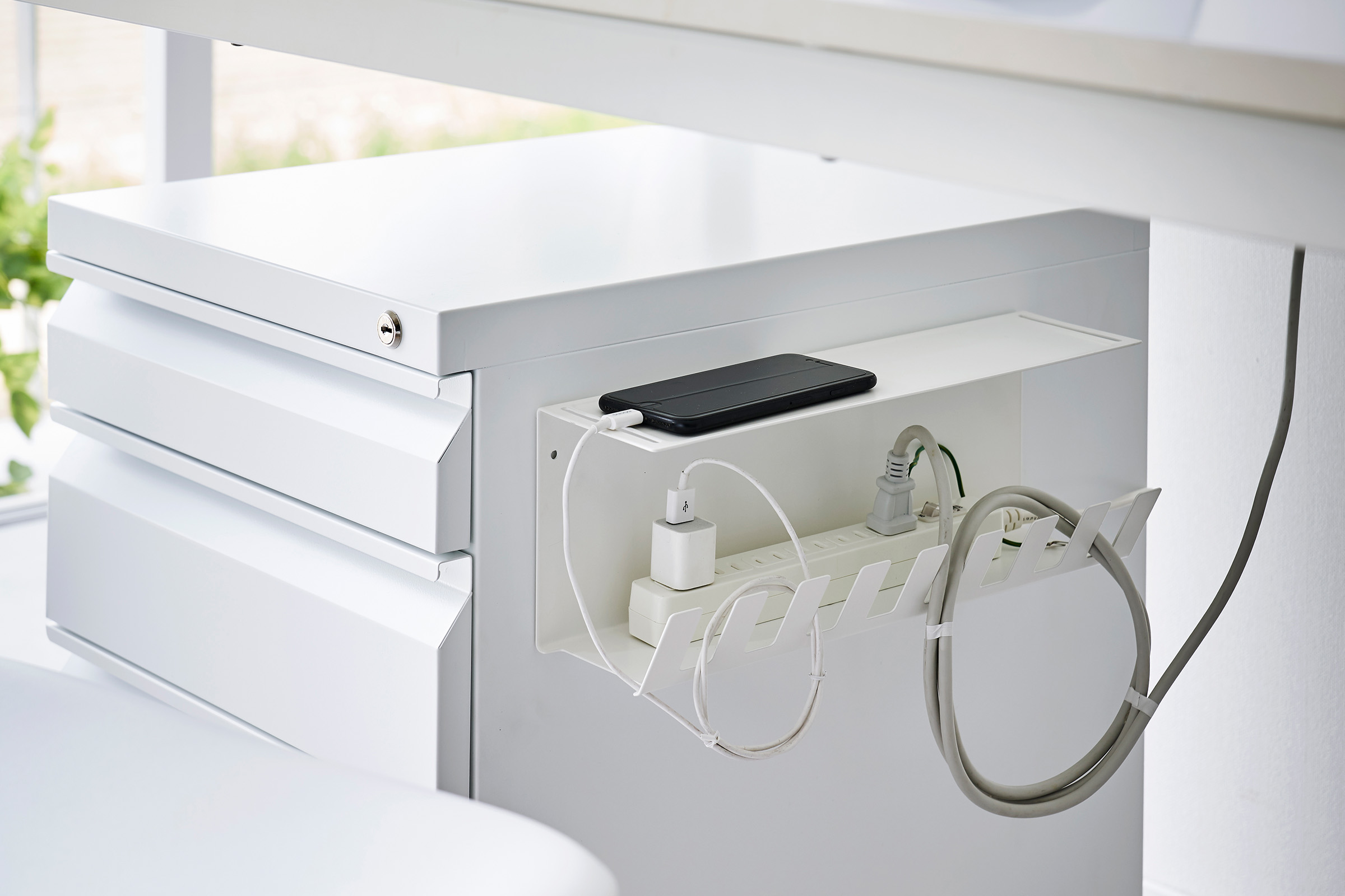 Magnetic Under-Desk Cable Organizer in white by Yamazaki Home mounted on the side of a file cabinet holding a power strip and phone.