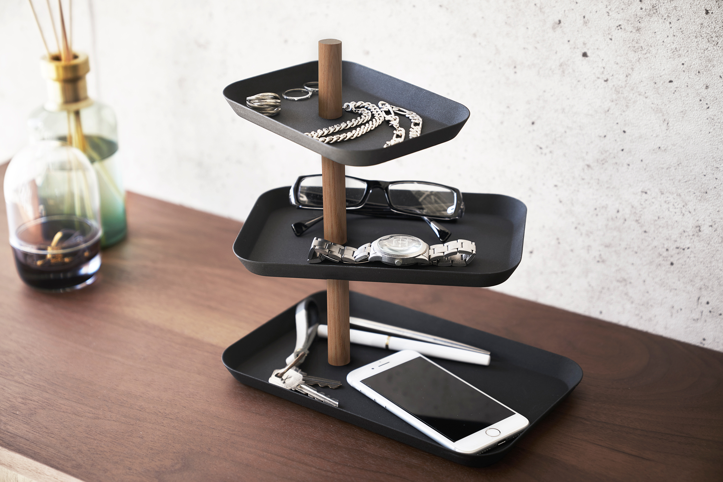 Yamazaki Home Black Rin Series 3-Tiered Accessory Tray with trays adjusted in a fan holding accessories such as a phone or glasses.