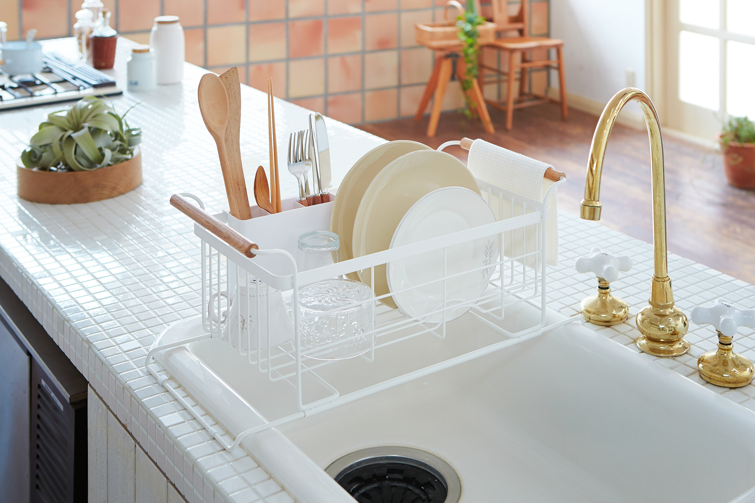 White Over-the-Sink Dish Rack holding plates, cups, bowls, and utensils on kitchen sink countertop by Yamazaki Home.
