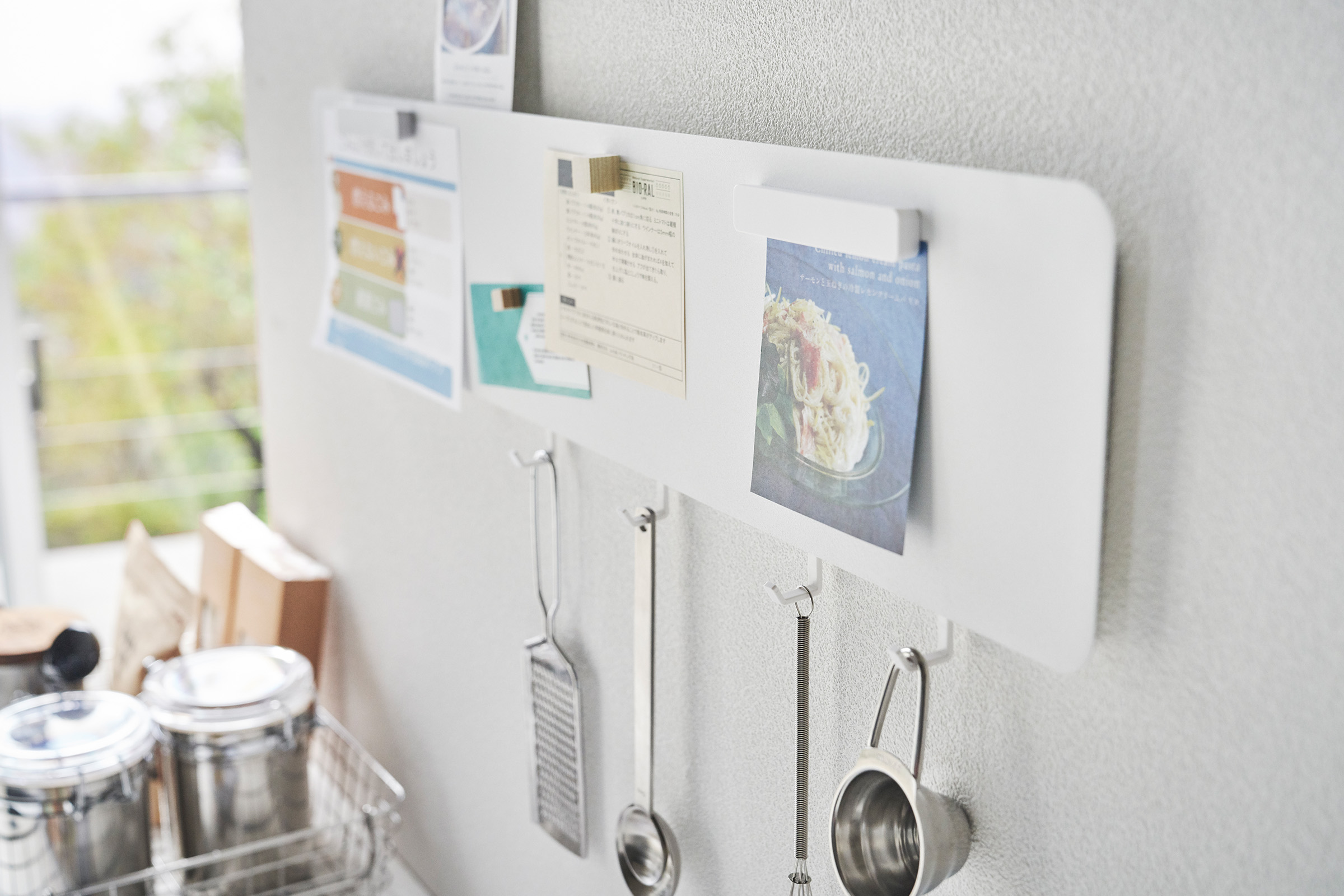 Magnetic Wall Panel in white by Yamazaki Home on a wall with various recipes tacked on and kitchen utensils hanging from bottom hooks.