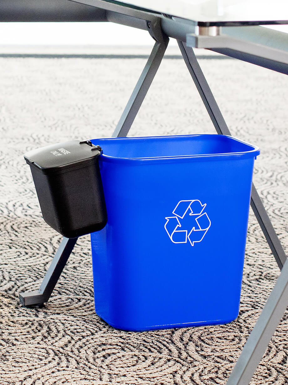 blue plastic deskside recycling container with black plastic hanging waste basket on the side sitting under a desk in a corporate office