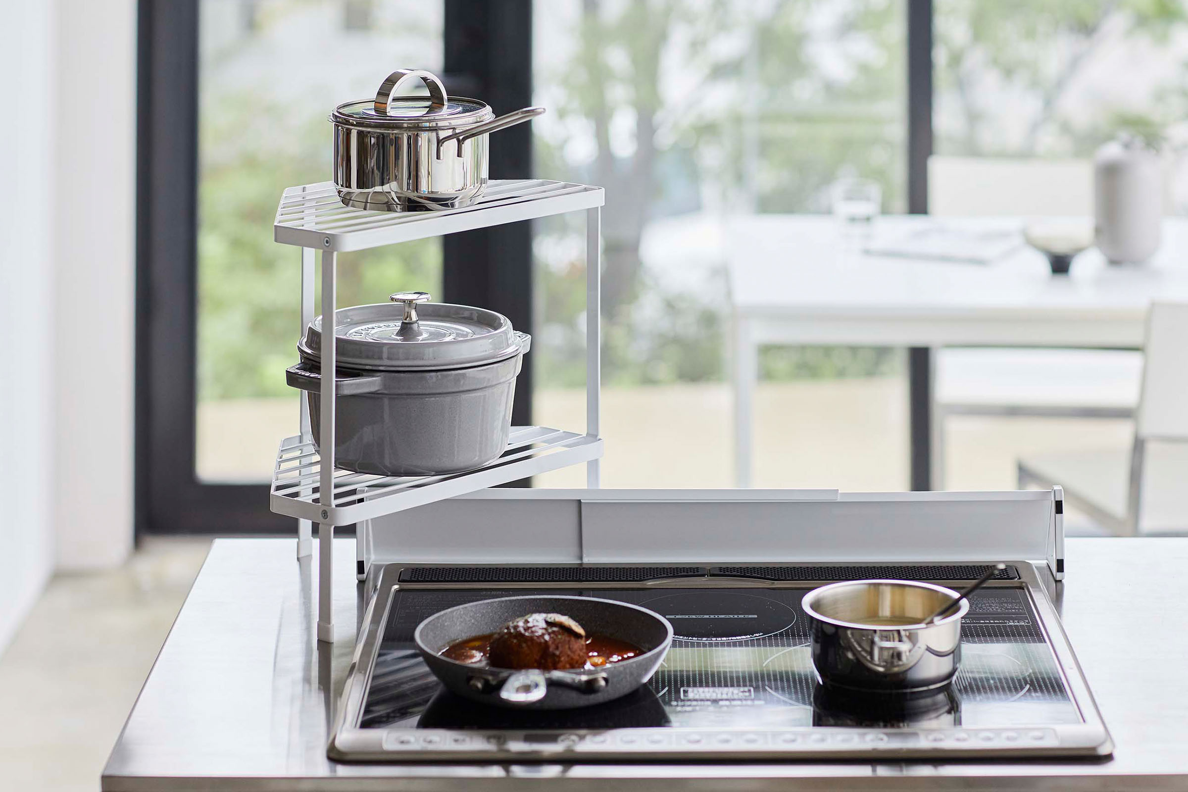 Front view of White Two-Tier Corner Riser holding pot and saucepan next to stovetop by Yamazaki Home.