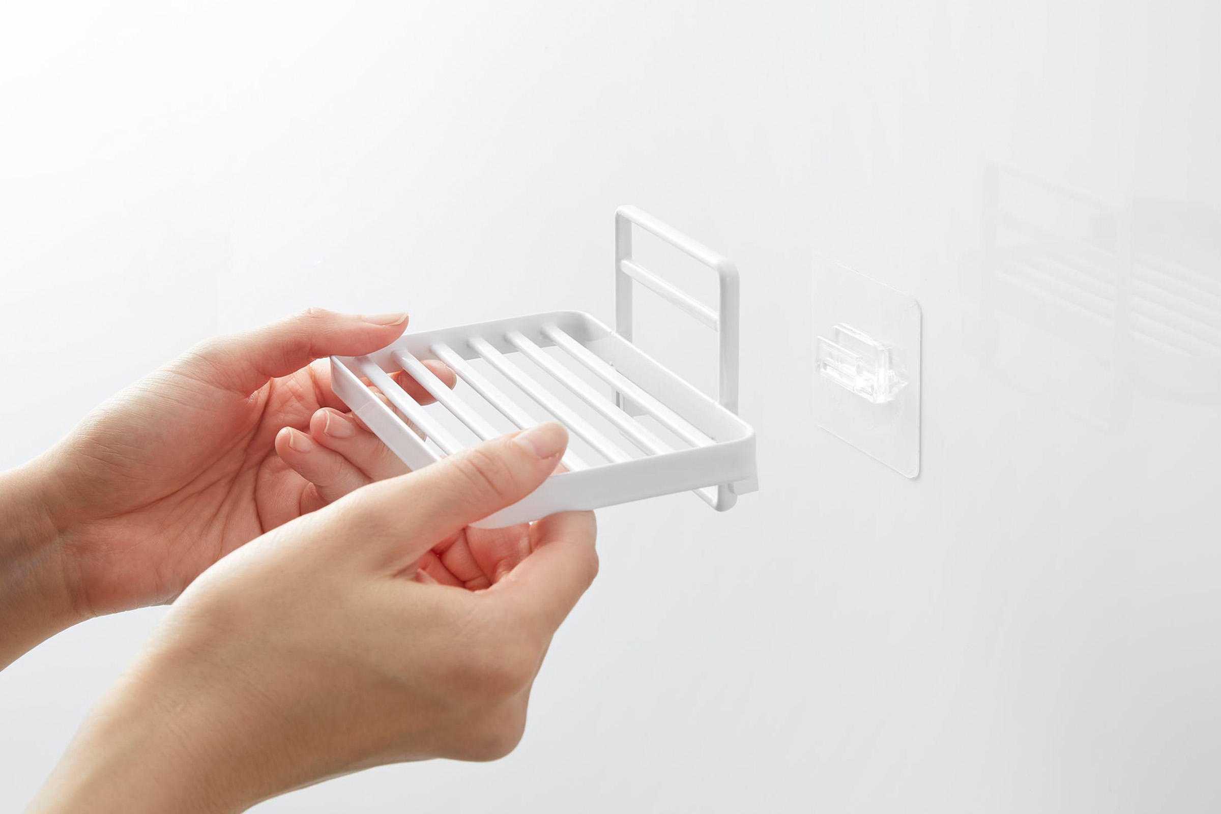 Model is shown installing a white steel soap tray to a translucent film hook placed on the wall. Soap tray has open slats for drainage.