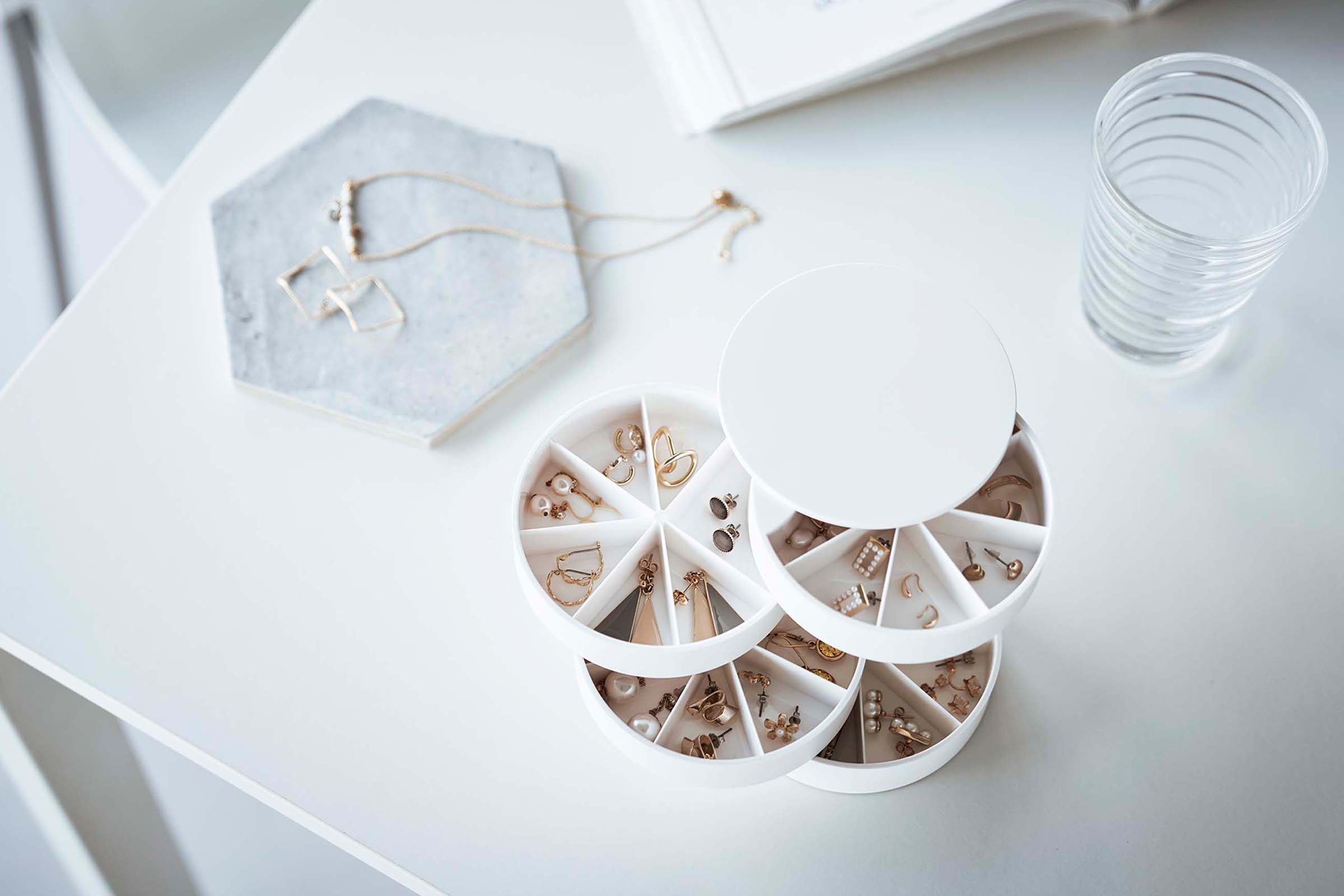 A bird’s-eye view of the corner of a white table. On the table are a cylinder-shaped white four-tier swivel accessory holder, a hexagon shaped catch-all dish and the corner of an opened book.