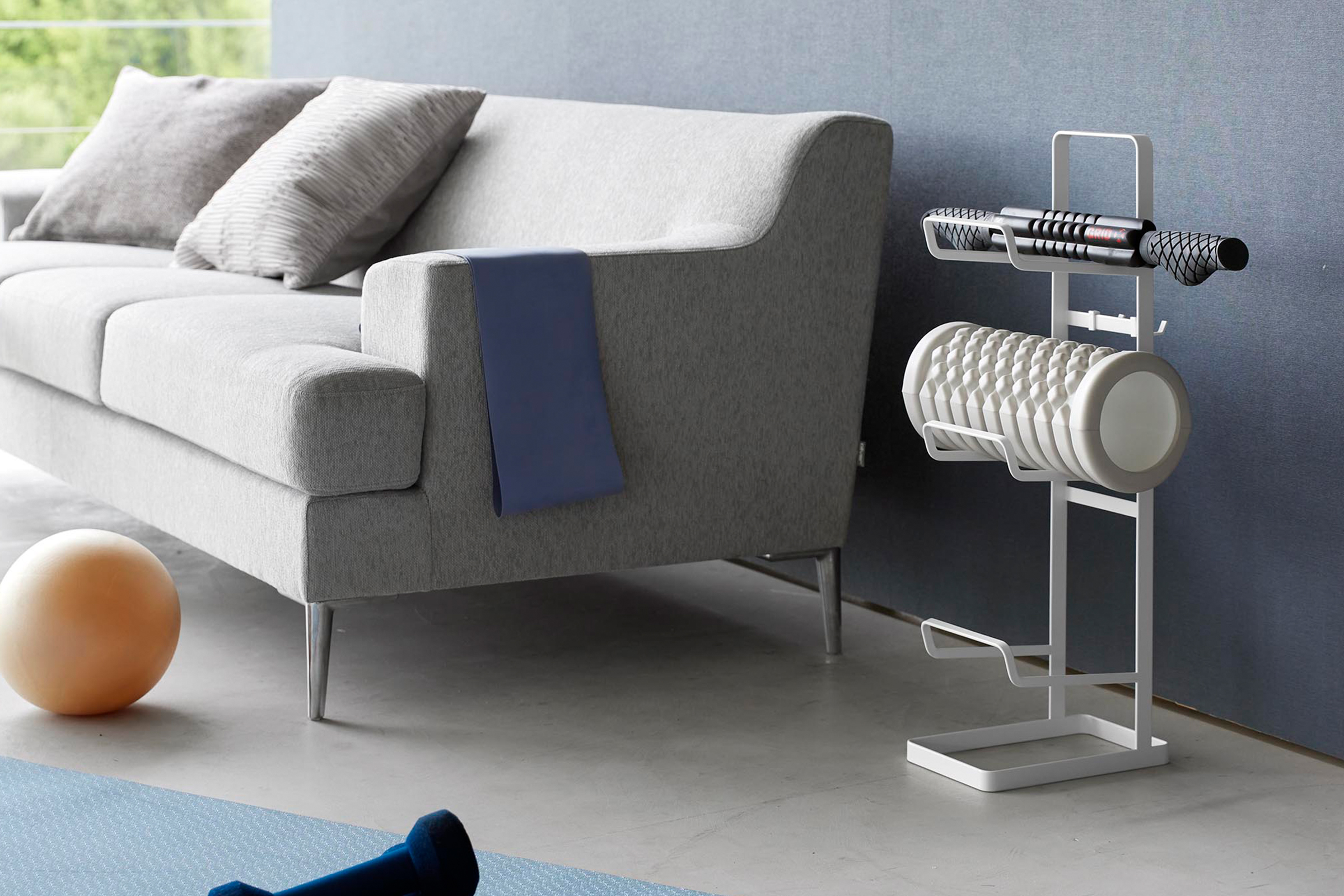 Wide view of Yamazaki Home White Fitness Equipment Storage Rack holding foam rollers next to sofa.