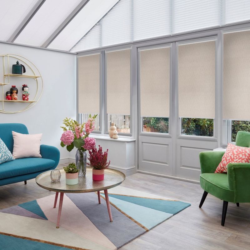 Beige roller blinds across tall conservatory windows and glazed doors, partially lowered to filter daylight; bright sunroom with skylight, teal sofa, green armchair, round coffee table and floral arrangements.