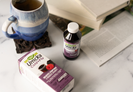 A bottle of Umcka Cold and Flu syrup sitting on a table next to some books and a coffee mug.