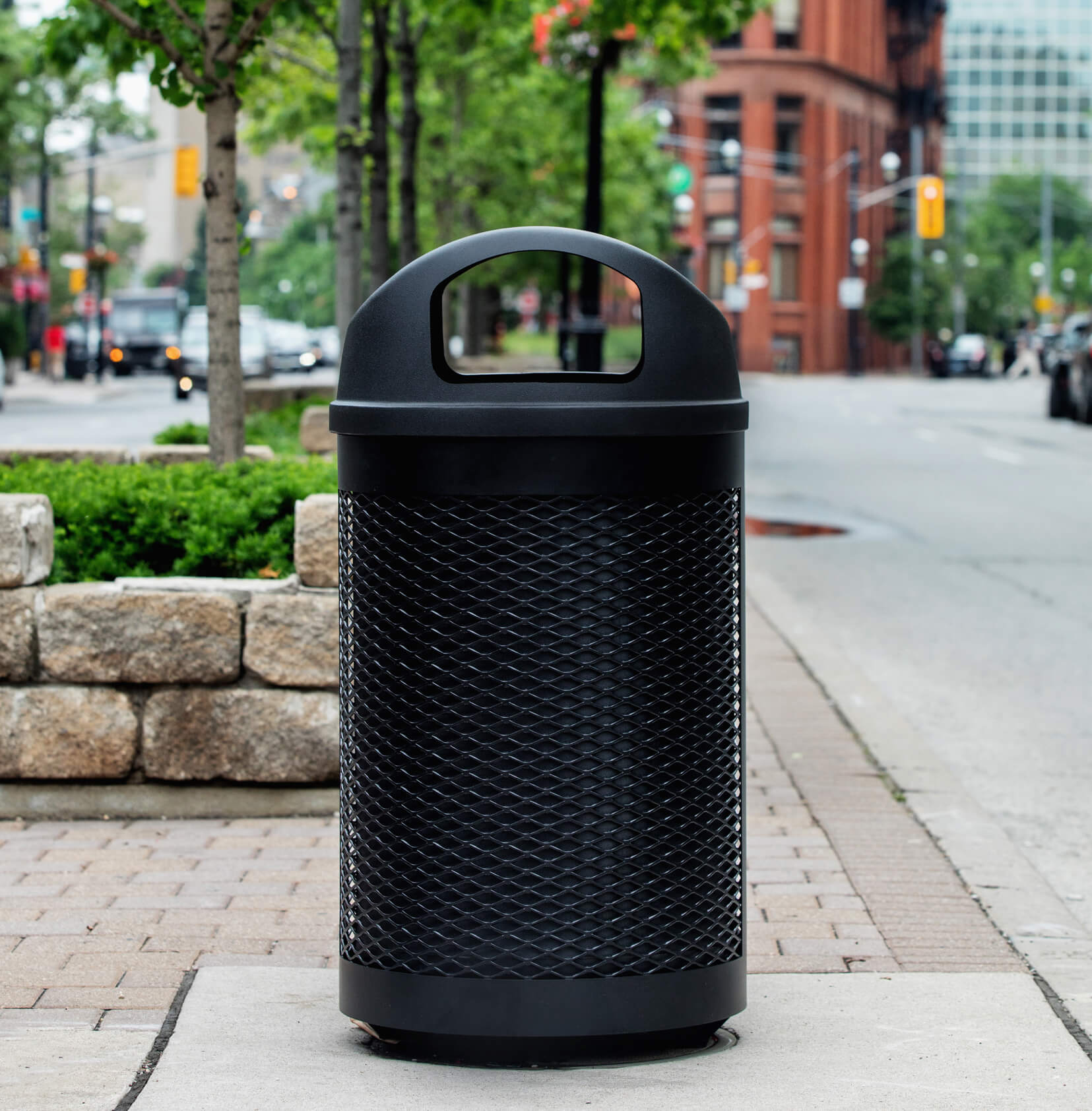 black powder coated steel waste container with a black plastic domed lid and liner on the side of a busy city street