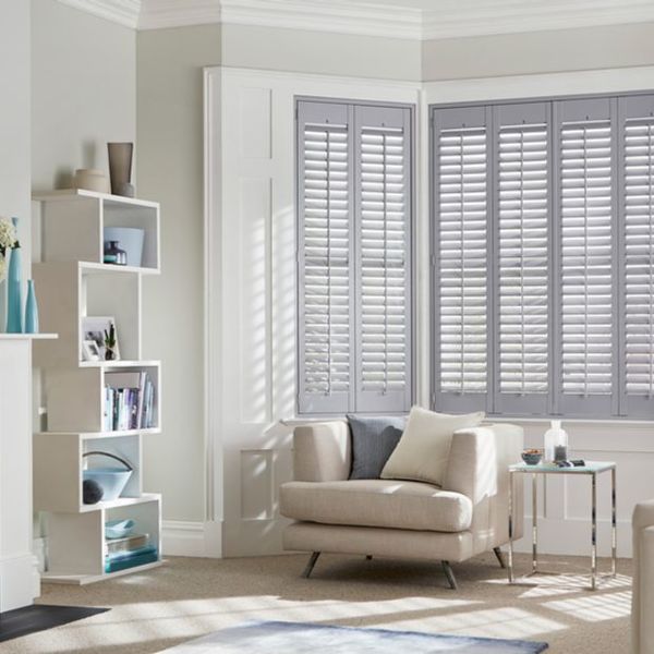 White plantation shutters on a bay window, closed with angled slats filtering sunlight into the room; neutral living room with beige armchair, throw pillows, open shelving and side table.