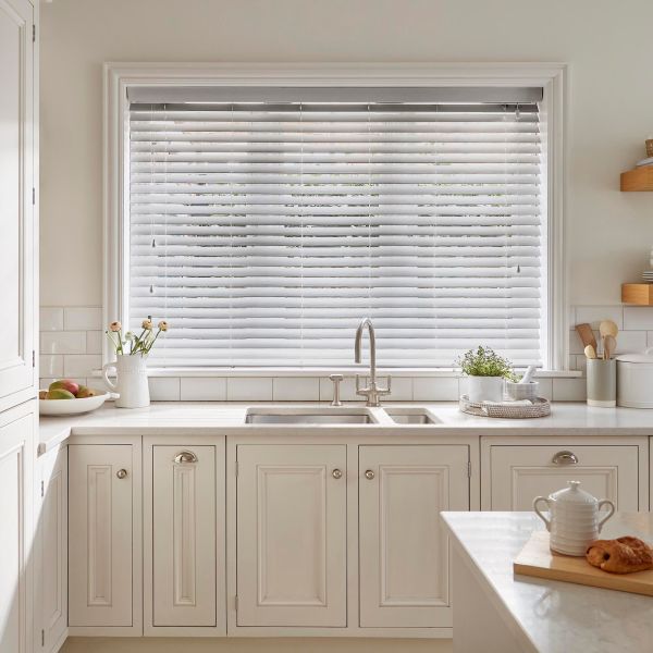 White horizontal Venetian blinds cover a large window, lowered with slats tilted to softly filter daylight; they sit above a farmhouse sink in a bright, neutral kitchen with cream cabinets.