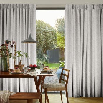 Pleated pale geometric curtains, hung full-length and parted centrally to frame a door, filtering soft daylight; a dining table with pancakes and pendant lamp sits before a leafy backyard.