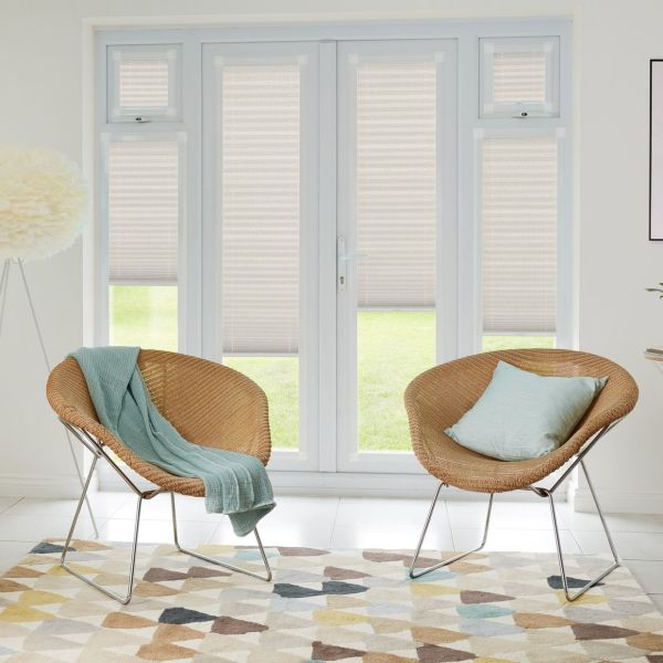 Beige pleated light‑filtering shades lowered over double French doors and narrow flanking windows, softly diffusing daylight; bright modern living room with two woven rattan chairs, pastel throw, and patterned rug.