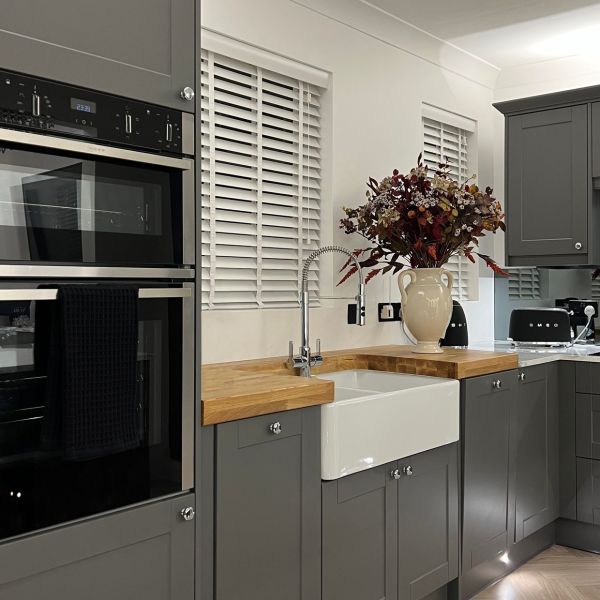 White horizontal slatted blinds over two windows, closed and softly filtering light; foreground farmhouse sink and wooden countertop within grey shaker cabinets, stainless ovens and a vase of autumn flowers.
SMEG
23:33