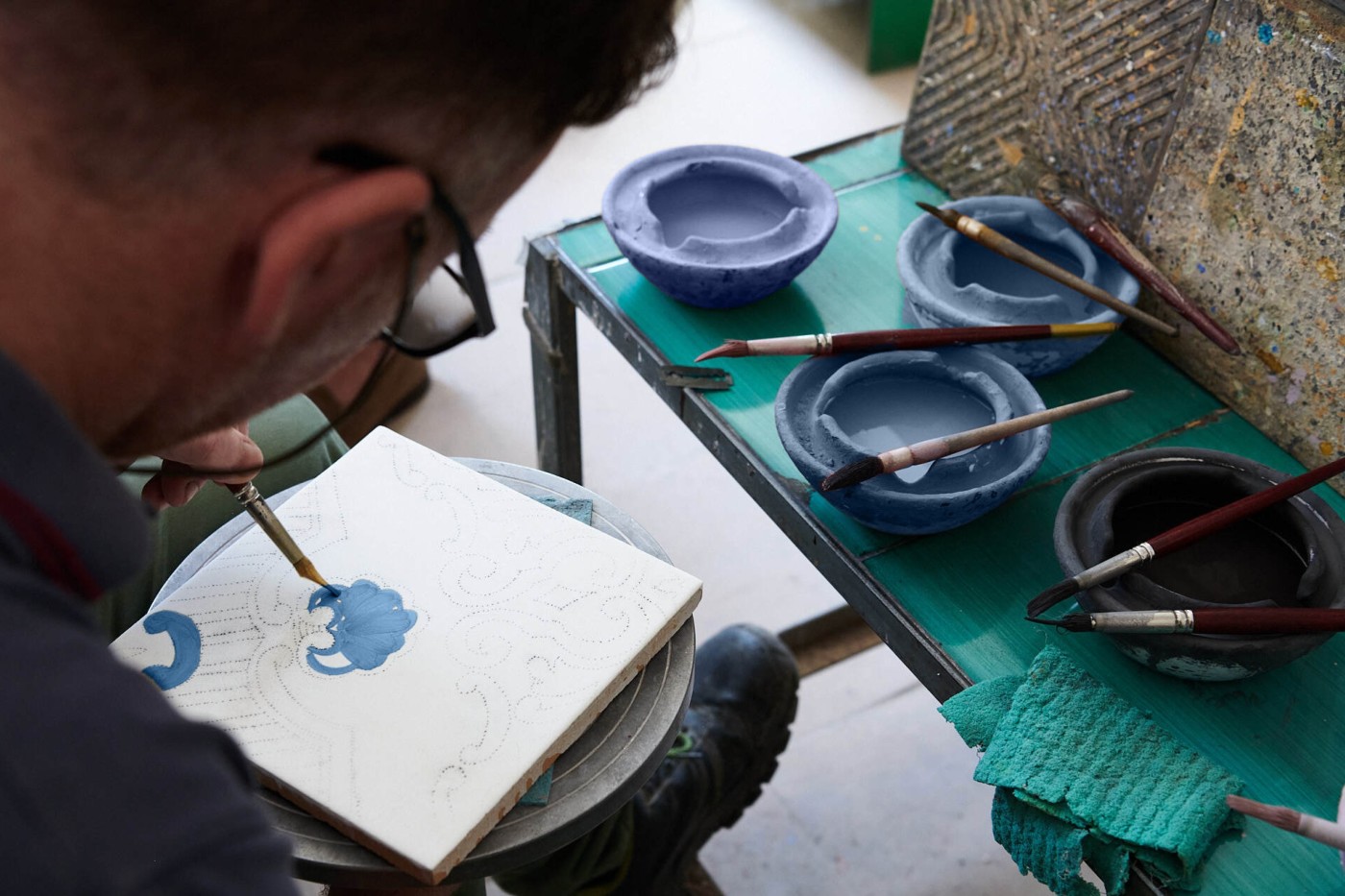 a man hand painting a blue design on a white tile.