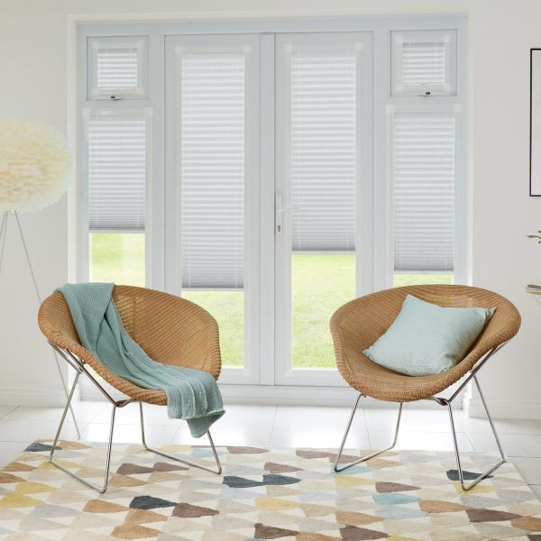 Pleated light-grey blinds on French doors and sidelights, partially lowered and filtering daylight, revealing lawn below; bright living room with two wicker chairs, pastel cushions, geometric rug and floor lamp.