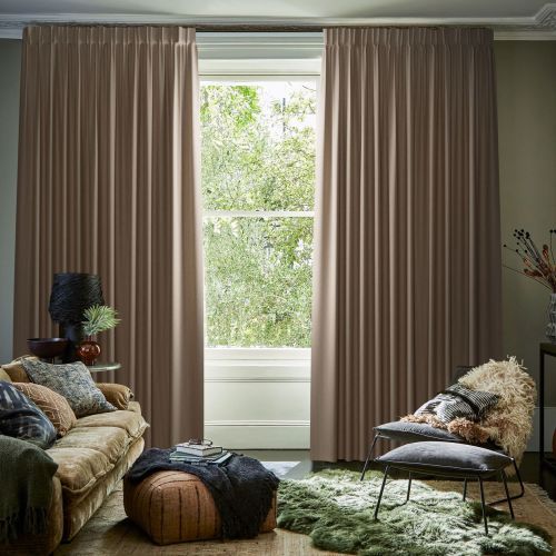 Full-length pleated beige curtains, hung on a ceiling track, mostly closed with a narrow central opening, framing and filtering daylight from a leafy garden visible through a living-room window.