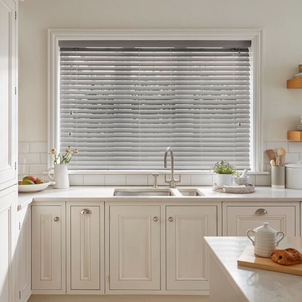 Horizontal Venetian blinds (grey slatted) covering a wide window, closed with slats slightly angled to filter daylight; above a white farmhouse kitchen sink, marble counters, potted herbs and utensils.