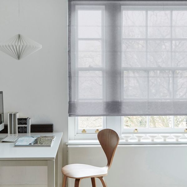 Sheer gray Roman shade (window dressing), lowered mid‑window and softly filtering daylight; simple white home office context with a wooden chair, desk, small pendant lamp and books.