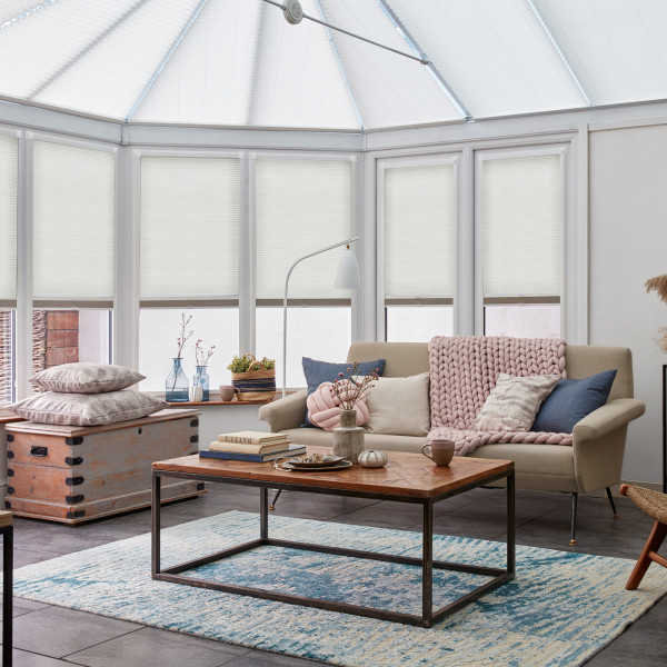 White pleated cellular blinds lowered over conservatory bay windows, softly diffusing daylight; beige sofa with knitted pink throw faces a wooden coffee table on blue rug in bright, airy sunroom.