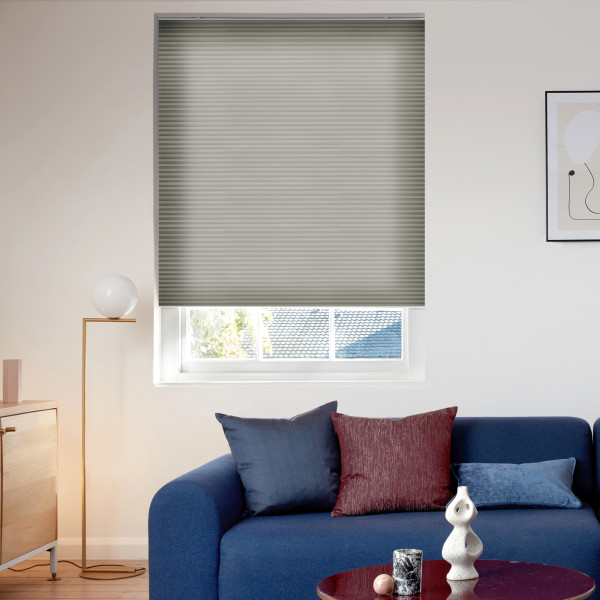 Gray pleated blind lowered over a window, filtering daylight; in a bright living room with blue sofa topped by navy and burgundy cushions, brass floor lamp and abstract wall art.