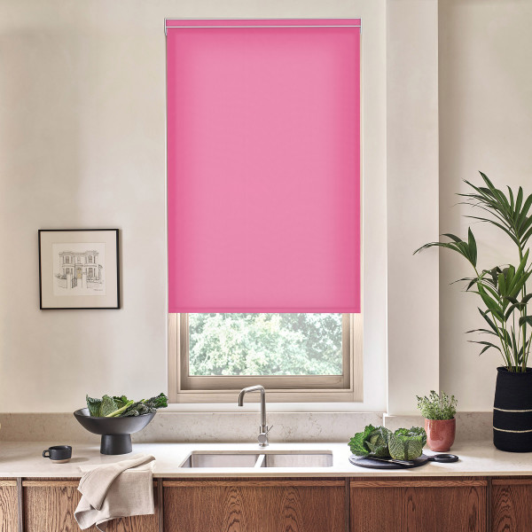 Pink roller blind covers a kitchen window, blocking sunlight; stainless faucet and double sink sit below on a marble countertop with potted plants, leafy greens, and framed sketch.