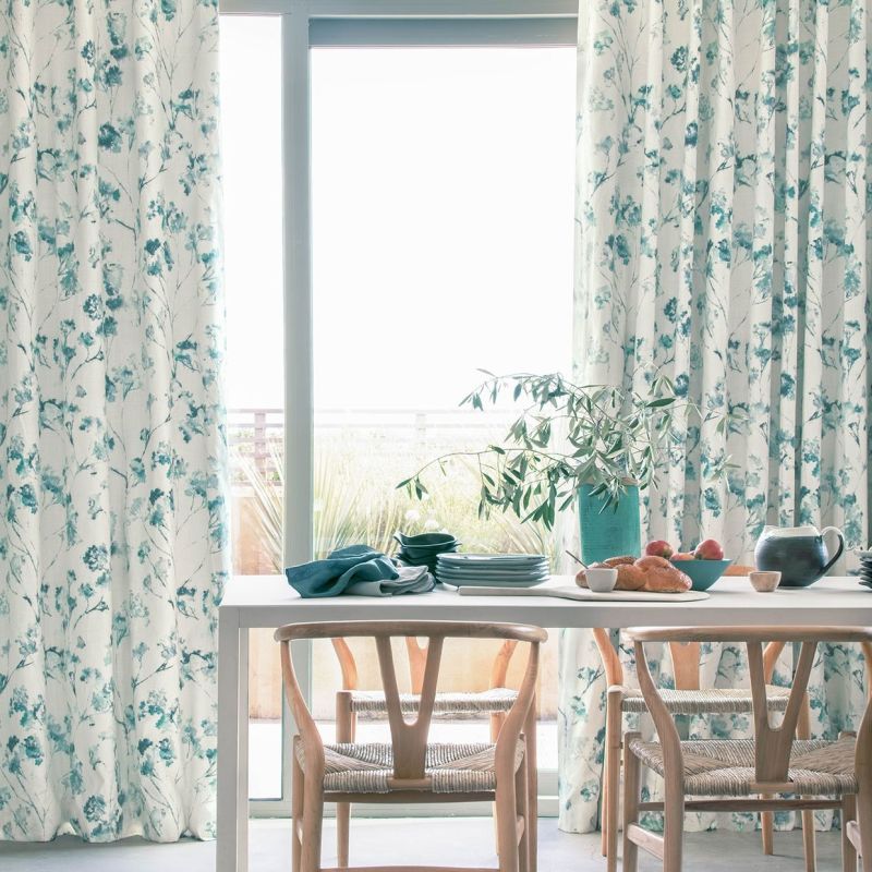 Floral-patterned, teal-and-white drapes, drawn aside to frame patio doors and filter bright daylight; dining table set with plates, fruit, bread and a vase, wooden chairs in a sunlit dining area.