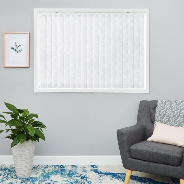 White fabric vertical blinds covering a centered window, closed and gently diffusing daylight; neutral living room with gray walls, potted plant, patterned rug, and a charcoal upholstered armchair.