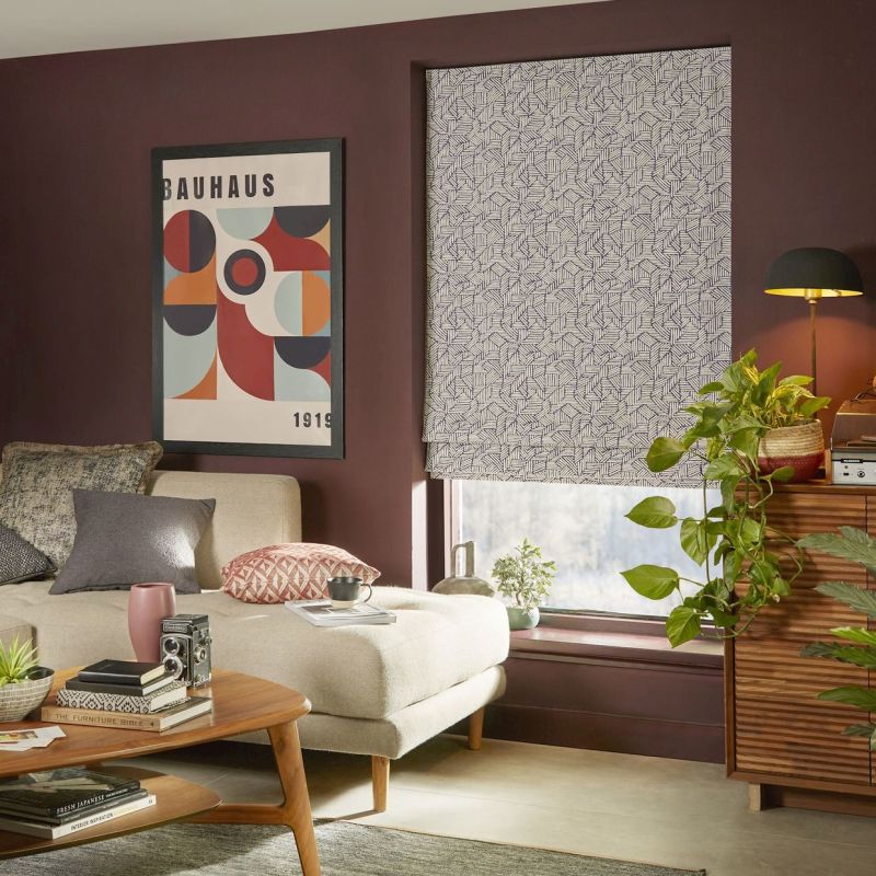 Roman blind lowered: white fabric with dense navy geometric line pattern, closed and softly diffusing light; in a plum-walled living room with a beige chaise, mid-century coffee table, plants, and poster.

BAUHAUS
1919