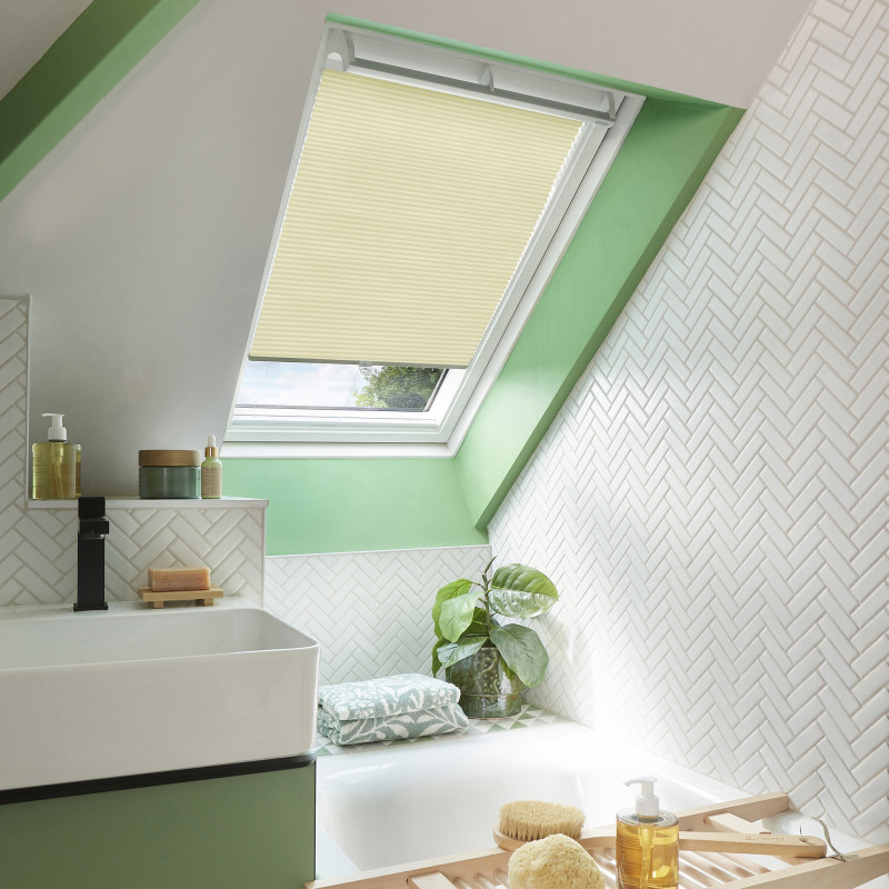 Cream pleated skylight blind lowered, filtering daylight; in a bright attic bathroom with mint-green window trim, white herringbone tiles, sink, bathtub, plant and toiletries on shelves.