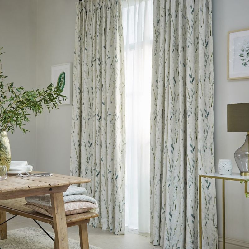 Floor-length pleated fabric curtains with green leaf print, hung on a window and closed over sheer panels, filtering daylight; set in a neutral dining nook with wooden table, plant, lamp.