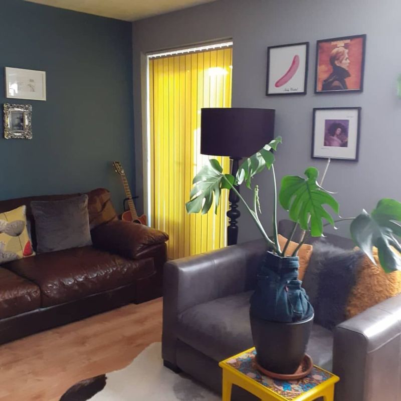 Yellow vertical blinds covering a sliding door, closed and filtering sunlight; in a living room with leather sofas, a lamp, potted monstera on a yellow table, framed art and guitar.
