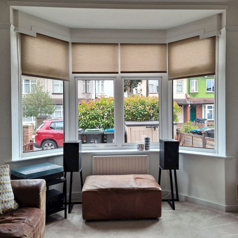 Beige roller blinds across a three-panel bay window, lowered and filtering daylight; white-trimmed living room with leather ottoman, speakers, radiator and street view of shrubs, parked cars and houses.