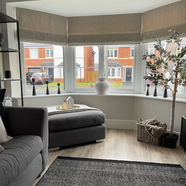 Beige fabric Roman blinds lowered across a threeâ€‘panel bay window, softly filtering daylight; decorative vase, candlesticks, potted tree and grey sofa/ottoman in a bright suburban living room.