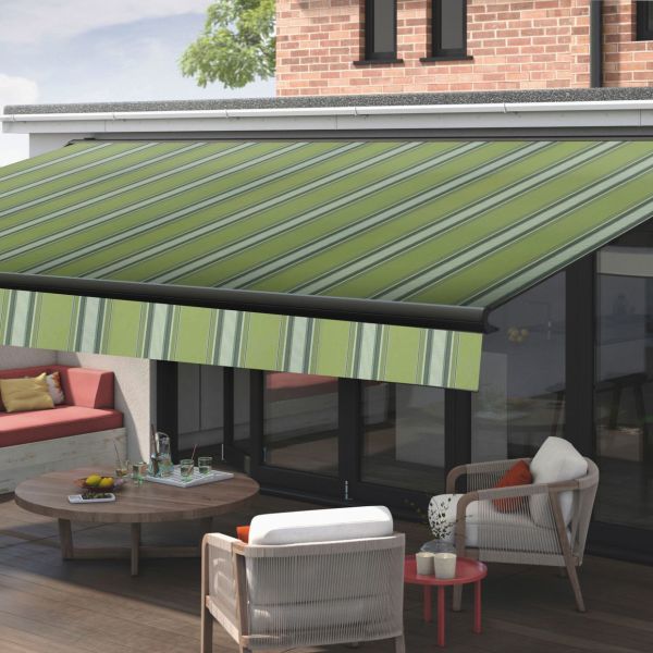 Green striped retractable awning, extended and angled downward, shading glass patio doors and a wooden deck; modern outdoor seating with chairs and low round table with drinks, brick upper wall, daylight.