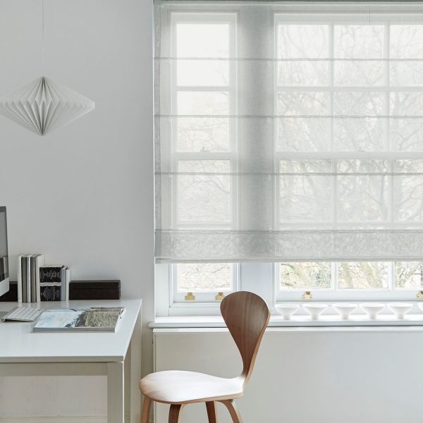 Sheer white Roman shade with horizontal folds, lowered over a multi-pane window, filtering soft daylight; minimalist home office with white desk, wooden chair, pendant lamp and bare tree branches outside.