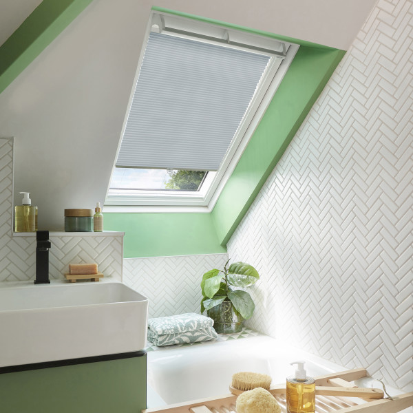 Pleated white cellular blind, lowered over a skylight window, filtering daylight. Bathroom attic space with green-painted trim, white herringbone tiles, sink, tub, potted plant and bath accessories.