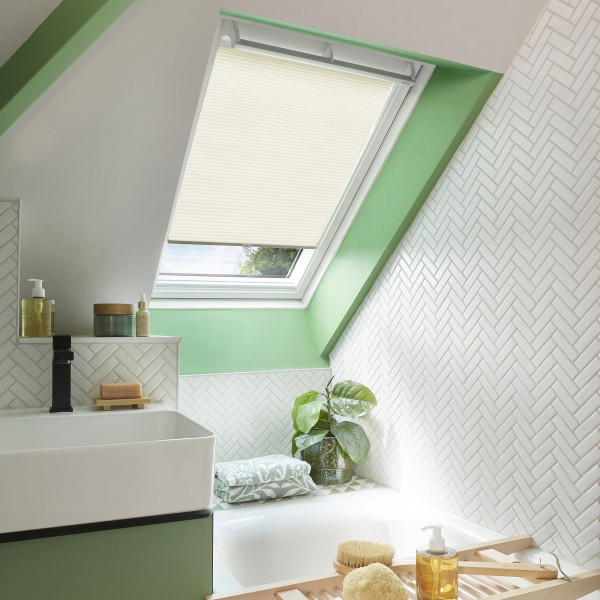 Cream pleated skylight blind, nearly lowered over an angled roof window, softly filtering daylight. Bathroom setting with green window surround, white herringbone tiles, bathtub, sink, plant and bath accessories.