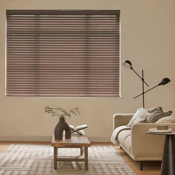 Horizontal blinds covering a wide window, slats closed with thin light lines; filtering muted daylight into a neutral living room with beige sofa, wooden table, rug, and floor lamp.