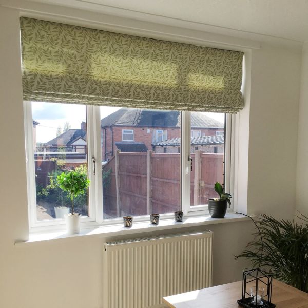Green leaf-patterned Roman blind, lowered partway and filtering sunlight; three-pane window with sill holding potted plants and candle holders; bright interior overlooking suburban houses and a wooden fence.