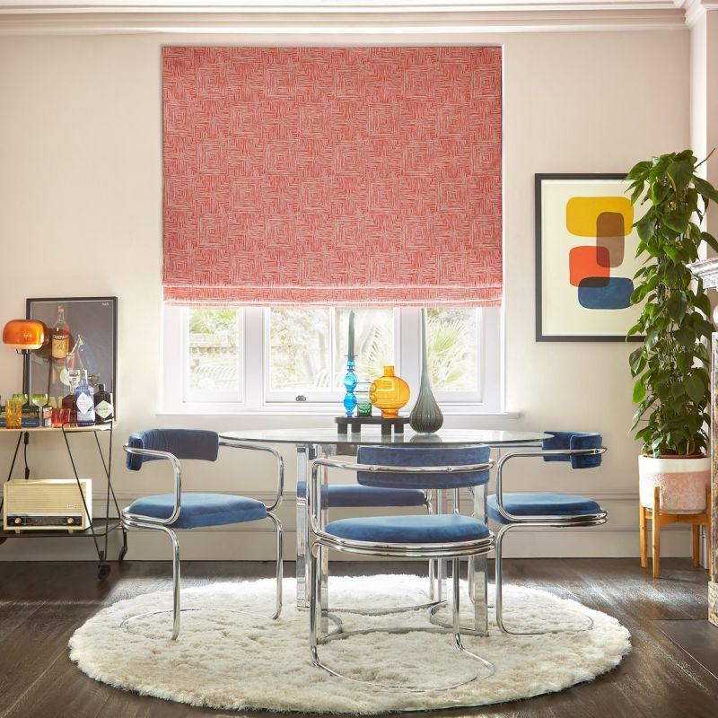 Red patterned Roman blind lowered over a threeâ€‘pane window, filtering daylight; glass dining table with chromeâ€‘framed blue chairs, round shag rug, potted plant and colorful vases; garden view. Campari