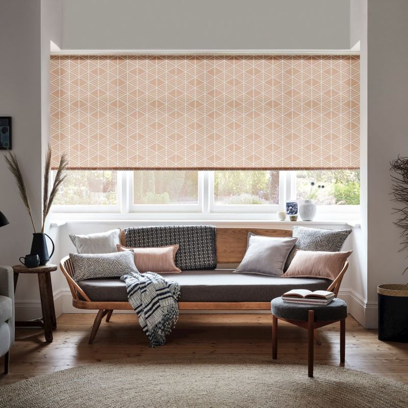 Peach geometric roller blind lowered over a bay window, filtering soft daylight through its lower gap; sits above a wood-framed sofa with cushions in a bright, cozy living room overlooking garden.