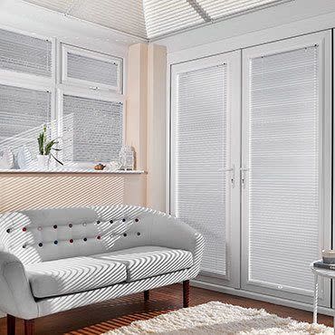 White pleated blinds covering high windows and French doors, closed and filtering sunlight into striped shadows across a curved two-seater sofa and sunlit living room with hardwood floor and rug.