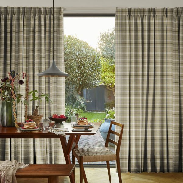 Beige plaid pleated curtains, mostly drawn with a central gap revealing the outside; filtering soft morning light over a wooden dining table set for breakfast, pendant lamp, and garden view.