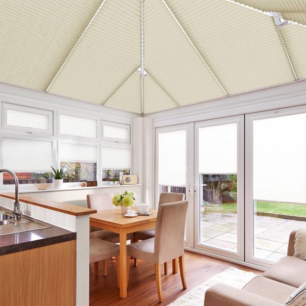 White pleated cellular blinds covering windows, patio doors and the pitched conservatory roof, closed and softly diffusing daylight; small dining area with wooden table, beige chairs, and garden view.