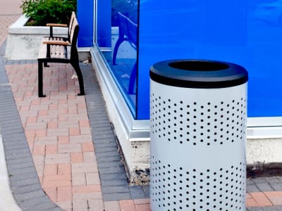 metal powder coated trash and recycling container with a perforated body and black plastic lid placed on a busy city sidewalk