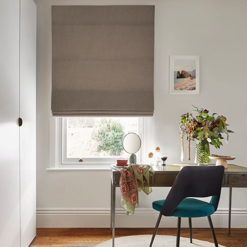 Taupe Roman blind, mostly lowered with soft folds, filtering daylight through a narrow gap; over a desk in a bright home office with vase of flowers, mirror and teal chair.
